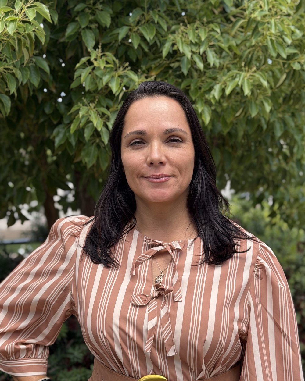 Adriana Lutti de vestido listrado, com a mão na cintura, diante de uma árvore