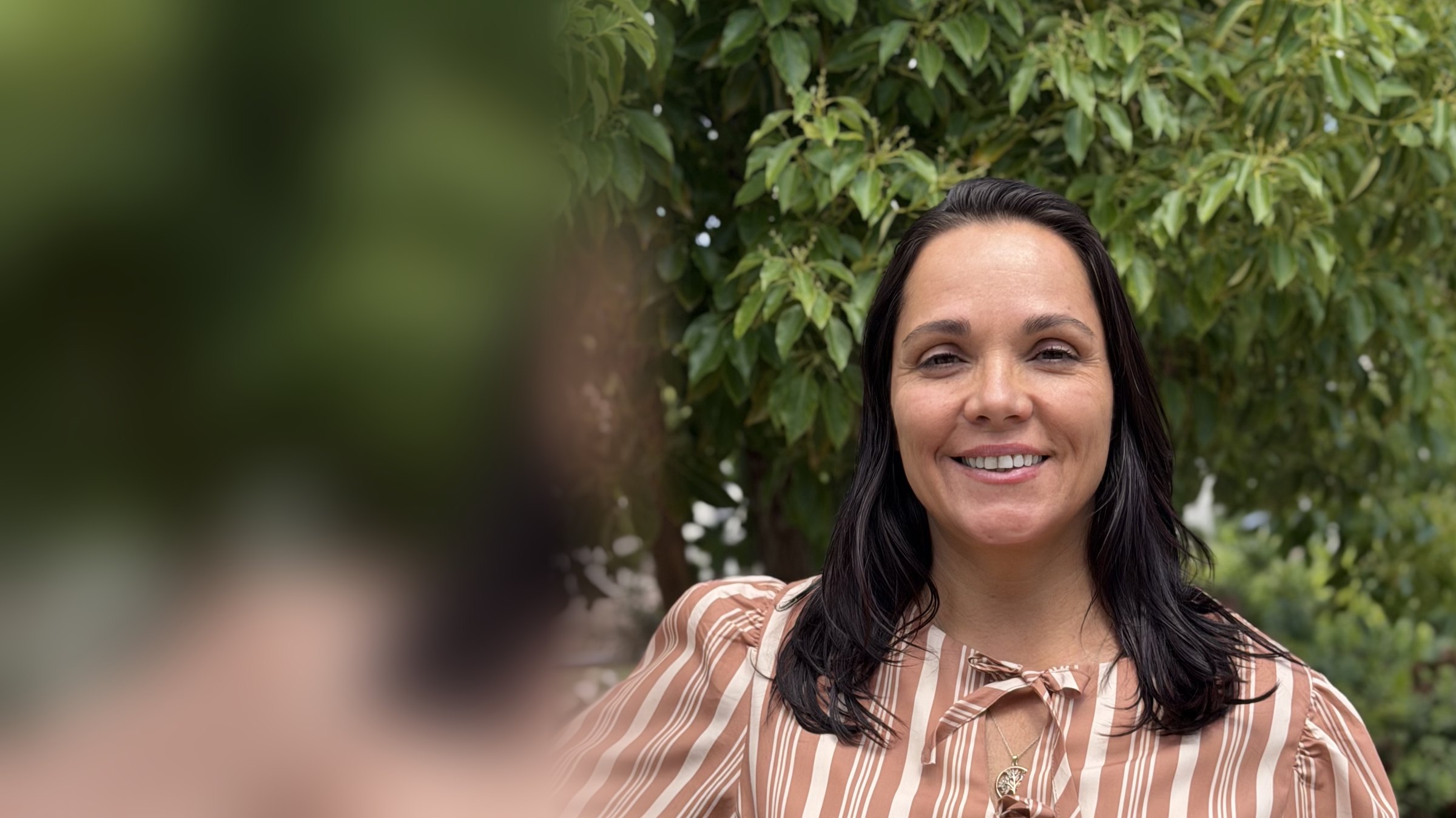 Adriana Lutti sorrindo ao ar livre, de vestido listrado, sob uma árvore